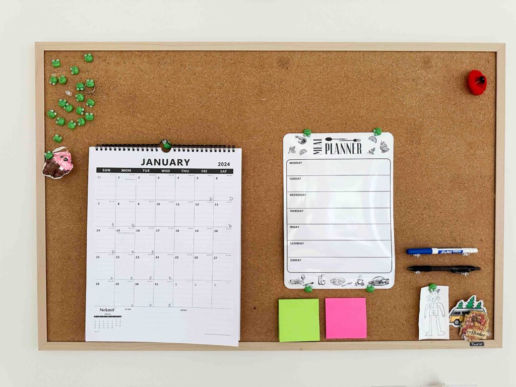 A madeover command center with pins, calendar, meal planner, post it notes, pens and two small pieces of children's artwork