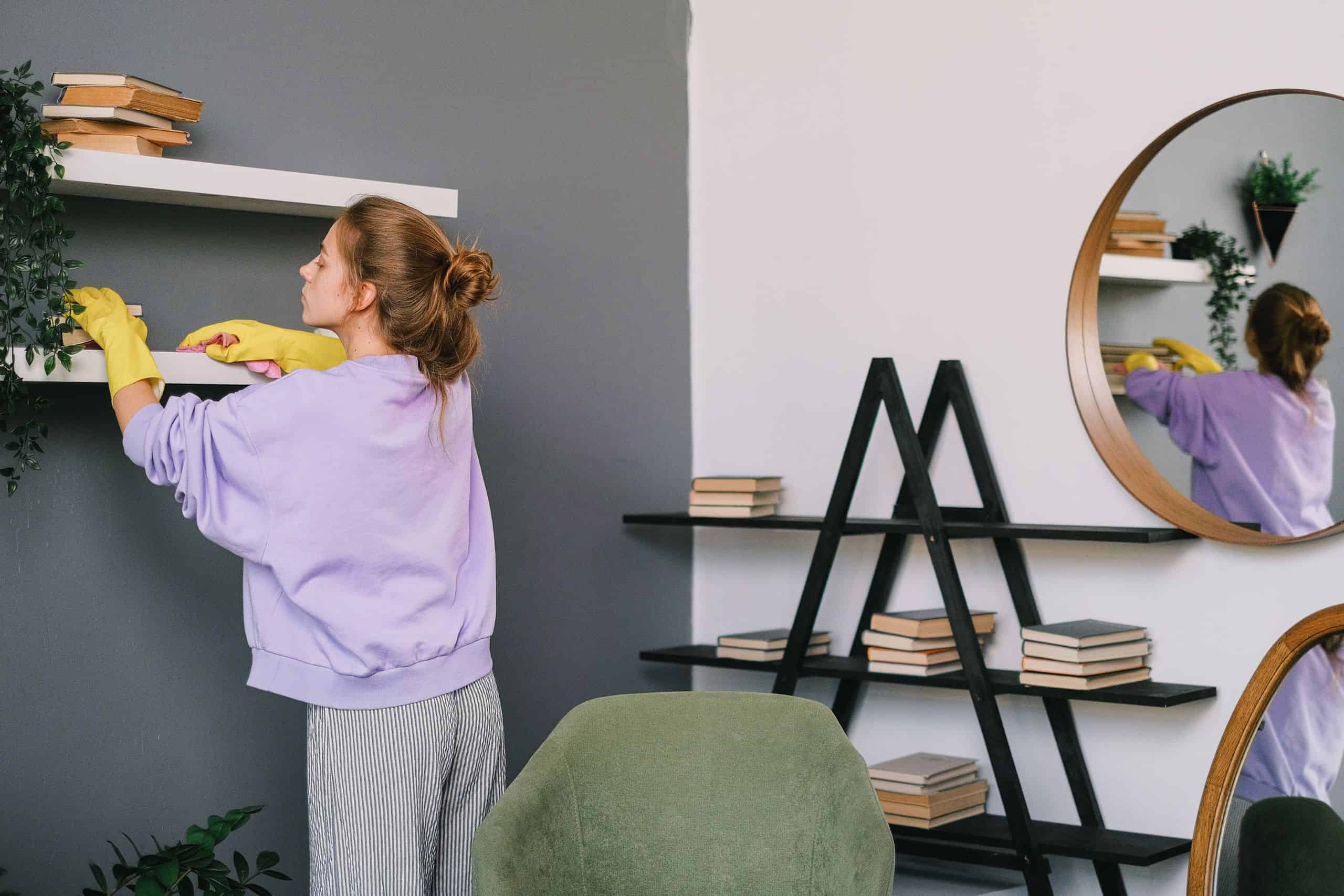 A woman cleans shelves in a stylish living room with mirrors and decor. PEXELS