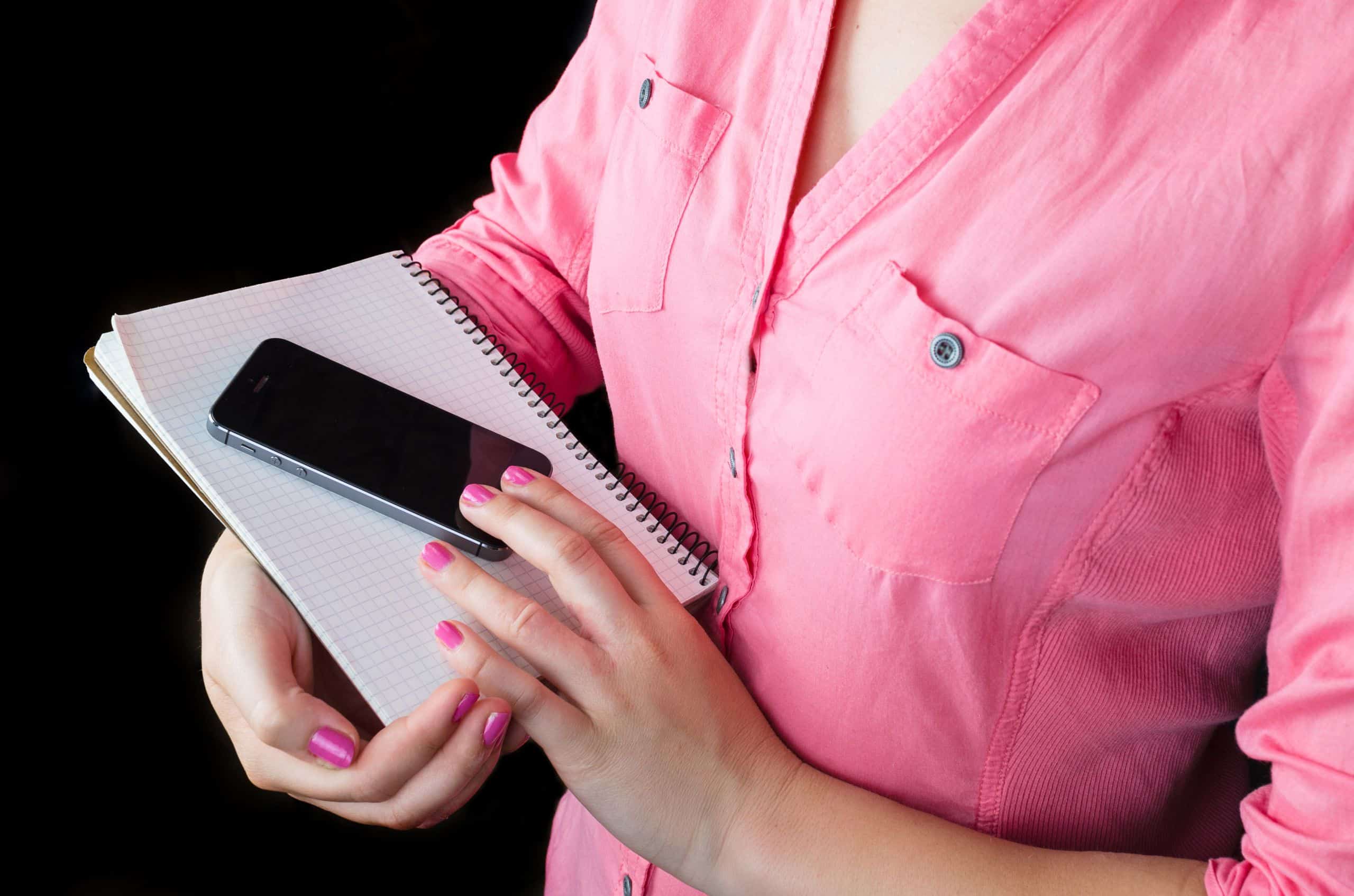 Close-up of a woman in a pink shirt holding a smartphone and notebook, focused on mobile technology and note-taking.