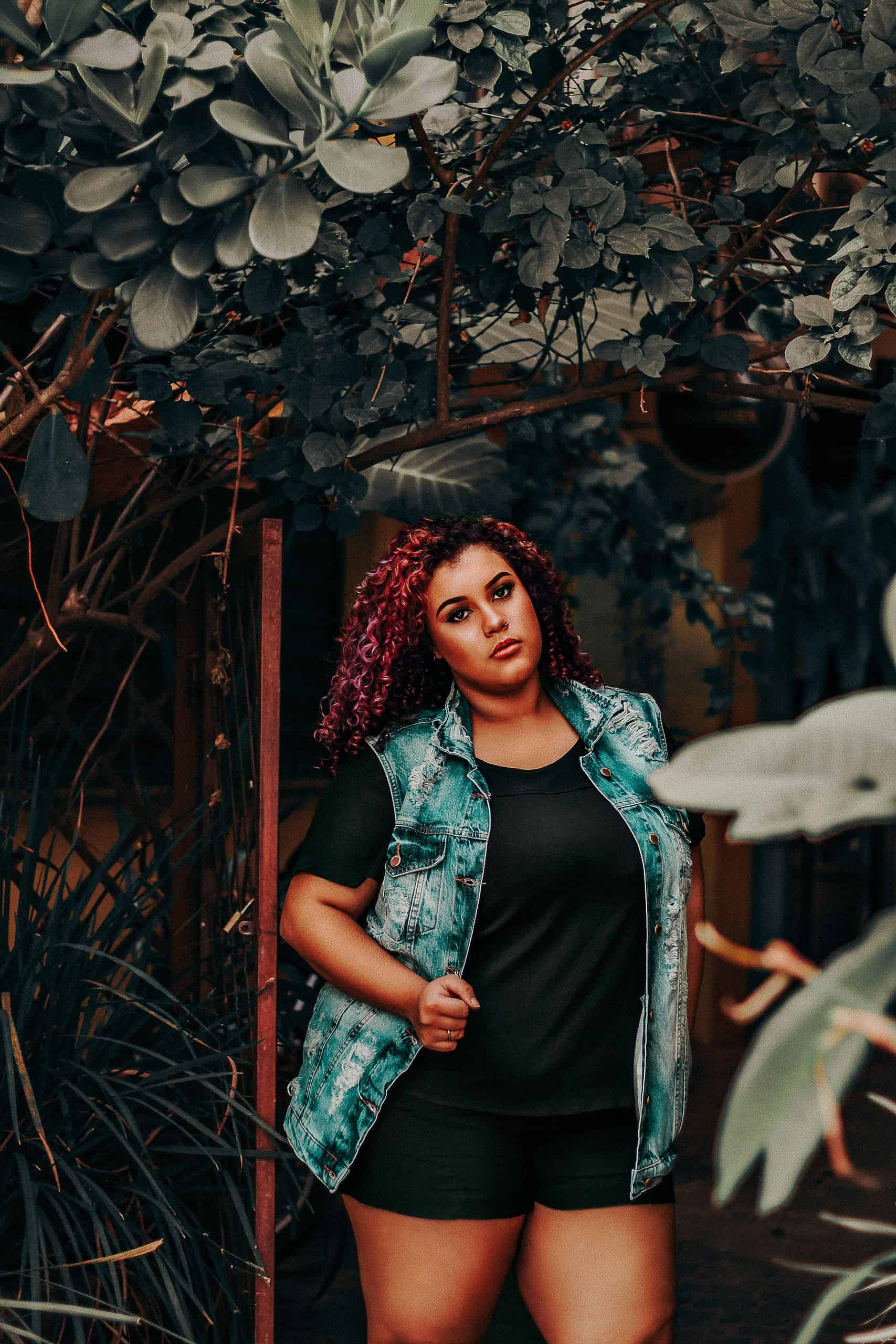 Portrait of a woman in a denim vest amidst lush greenery, exuding confidence and style.