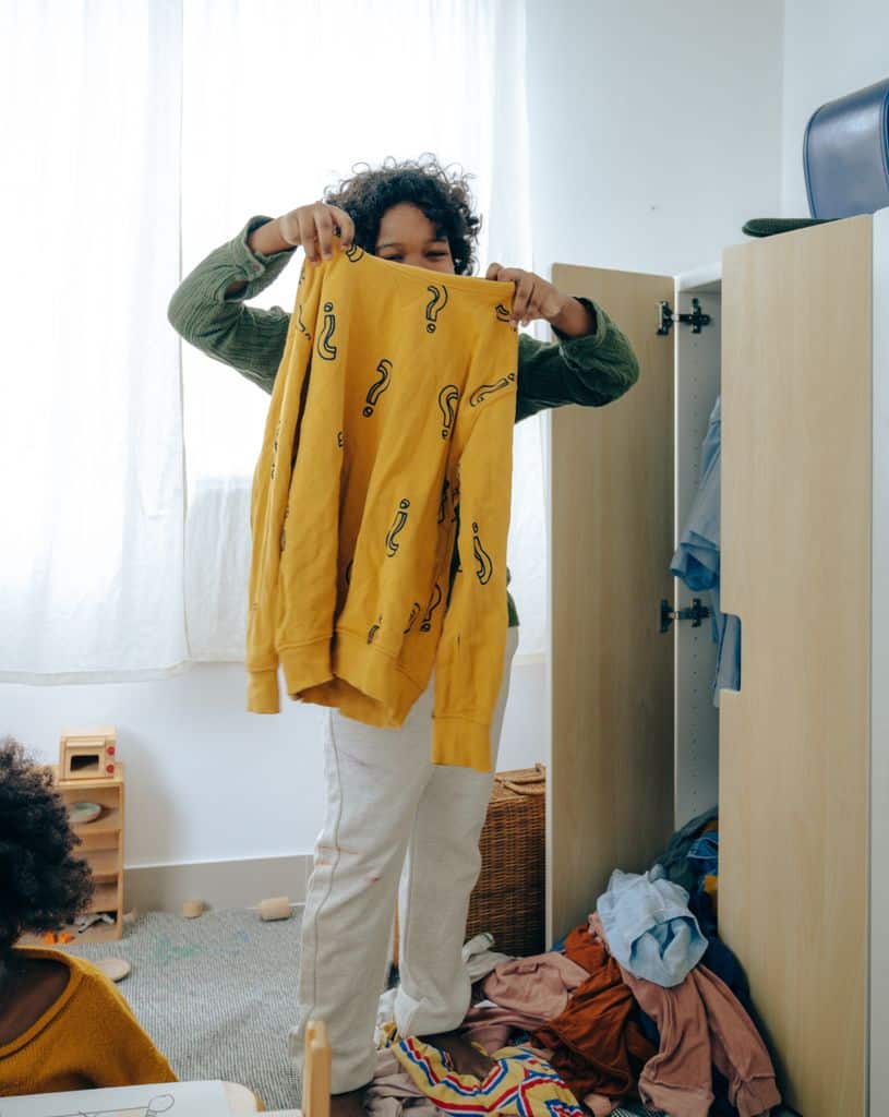 Unrecognizable African American teen girl in casual wear holding yellow sweater while sorting clothes in wardrobe in room. PEXELS